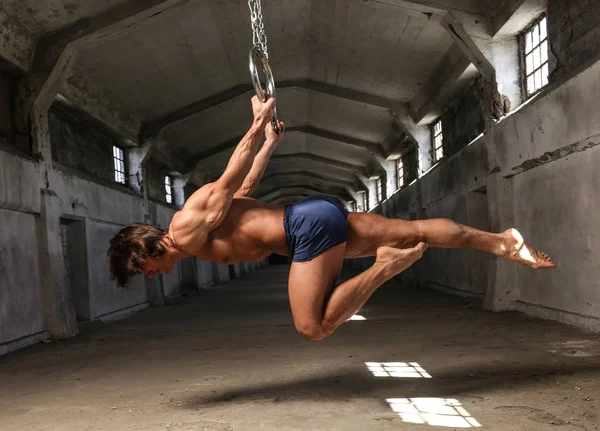 Um atleta profissional com corpo muscular fazendo exercício de ginástica em anéis, mostra a suspensão horizontal no edifício industrial abandonado — Fotografia de Stock