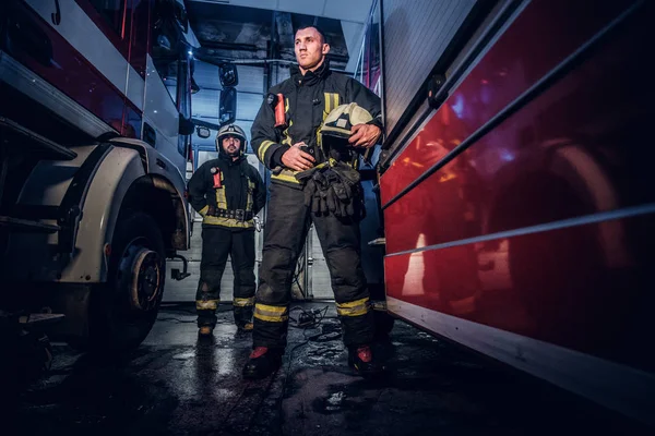 Két bátor tűzoltók a védő egységes állandó között két tűz motorok a garázsban a tűzoltóság teljes hosszúságú portréja — Stock Fotó