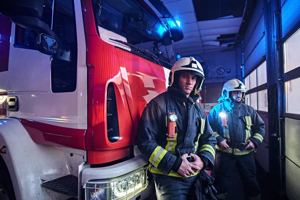 Zwei Feuerwehrmänner in Schutzuniform stehen neben einem Feuerwehrauto in einer Garage einer Feuerwehr. — Stockfoto