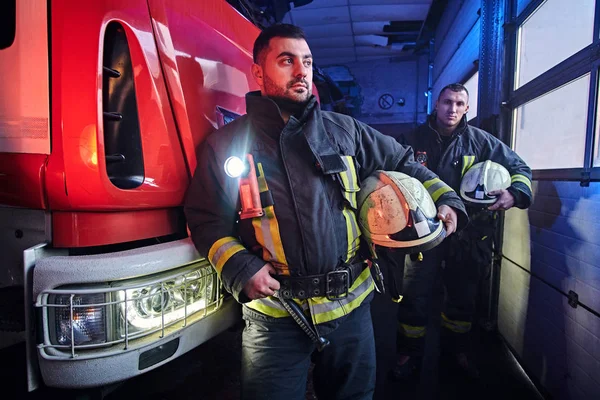 Dos bomberos con uniforme protector de pie junto a un camión de bomberos en un garaje de un departamento de bomberos . — Foto de Stock