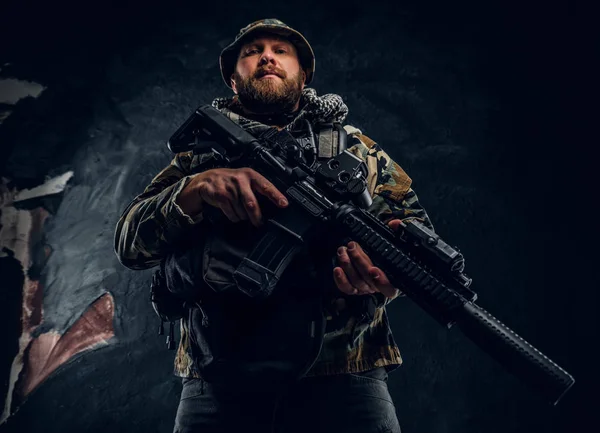 Żołnierz sił specjalnych w wojskowym mundurze zakamuflowane trzyma karabin szturmowy. Studio fotografii ścianę teksturowane ciemne — Zdjęcie stockowe