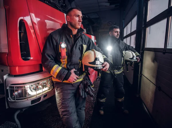 Dva hasiči nosí ochranné jednotný stát vedle hasičské auto v garáži hasiči. — Stock fotografie