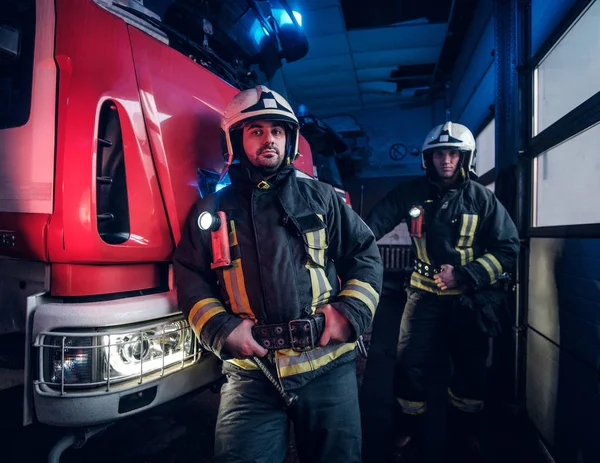Dva hasiči nosí ochranné jednotný stát vedle hasičské auto v garáži hasiči. — Stock fotografie
