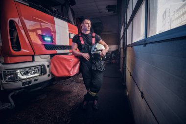 Young fireman holding a jacket and protective helmet in a garage of a fire department, leaning on a fire engine and looking outside clipart