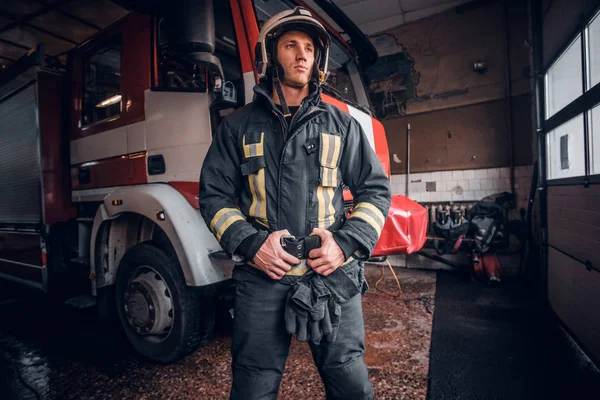 Joven bombero con uniforme protector de pie junto a un camión de bomberos en un garaje de un departamento de bomberos — Foto de Stock