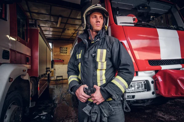 Fiatal tűzoltó visel védő egységes mellette egy tűzoltóautó, egy garázs, egy tűzoltóság — Stock Fotó