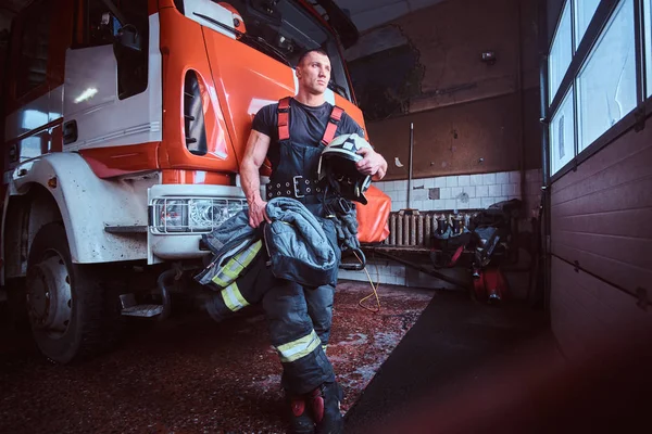 Joven bombero sosteniendo una chaqueta y un casco protector en un garaje de un departamento de bomberos, apoyado en un camión de bomberos y mirando hacia el exterior — Foto de Stock