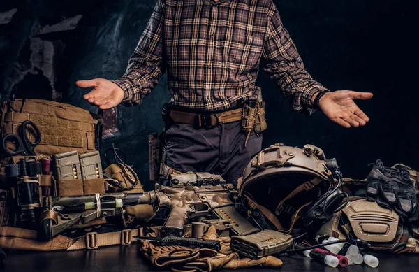 Un uomo con una camicia a scacchi che mostra la sua uniforme militare e la sua attrezzatura. Moderna attrezzatura delle forze speciali. Studio foto contro un muro scuro strutturato — Foto Stock