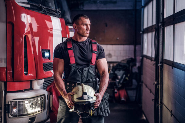 Muscular fireman holding a protective helmet in a garage of a fire department