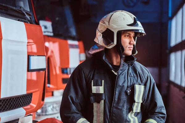 Fiatal tűzoltó visel védő egységes mellette egy tűzoltóautó, egy garázs, egy tűzoltóság — Stock Fotó