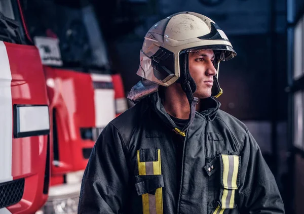 Young fireman wearing protective uniform standing next to a fire engine in a garage of a fire department — Stock Photo, Image