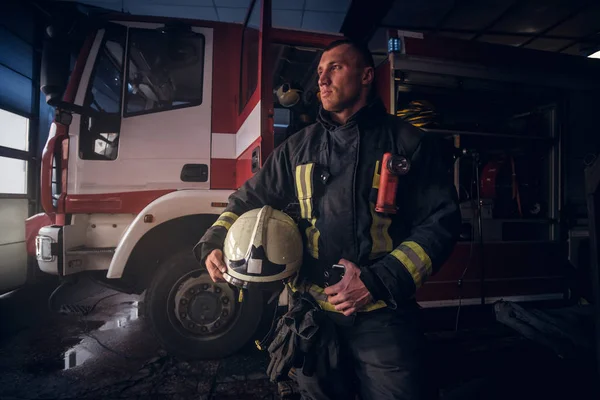 Fireman wearing uniform holding a helmet and looking sideways while standing near a fire truck in a garage of a fire department — Stock Photo, Image