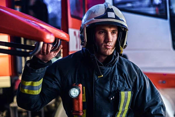 Retrato de un guapo bombero vestido con un uniforme protector con linterna incluido de pie en un garaje de bomberos — Foto de Stock