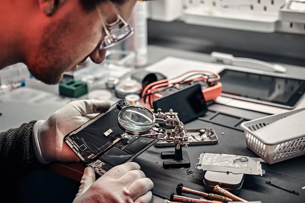 Techniker untersucht in einer modernen Werkstatt mit einer Lupe sorgfältig die Innenteile des Smartphones — Stockfoto
