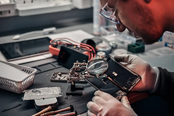 O técnico usa uma lupa para inspecionar cuidadosamente as partes internas do smartphone em uma oficina de reparo moderna — Fotografia de Stock