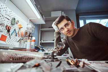 Düşünceli bir bakışla bir kameraya bakarak elektronik teknisyeni gözlük, bir tamir dükkanında bir masa üzerine eğilerek içinde