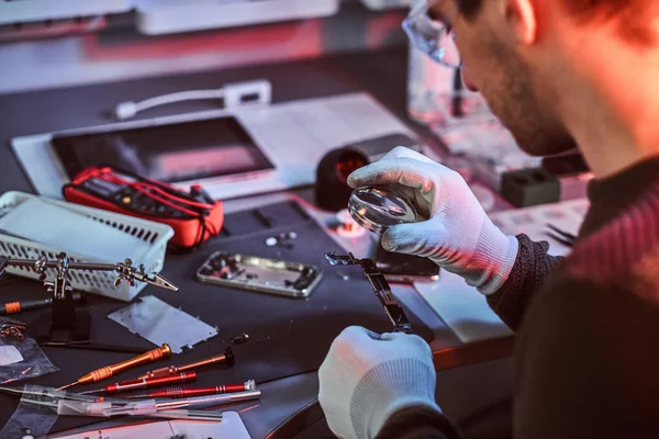 Der Elektroniktechniker, der eine kaputte Tablette repariert, untersucht den Chip der Tablette sorgfältig mit einer Lupe. Beleuchtung mit rotem und blauem Licht — Stockfoto