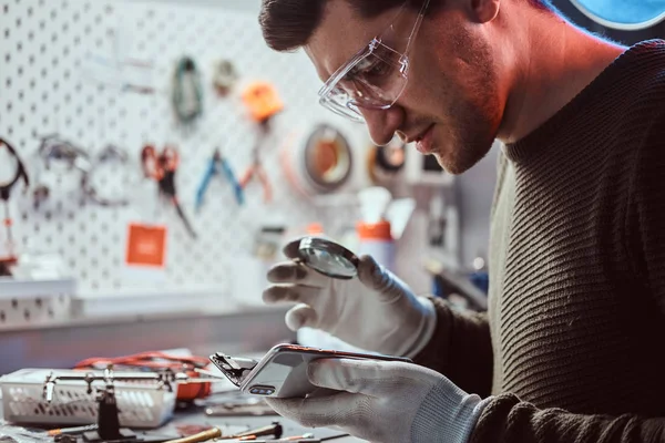 Technicien utilise une loupe pour inspecter soigneusement les parties internes du smartphone dans un atelier de réparation moderne — Photo