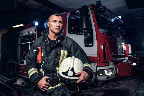 Bombero vistiendo uniforme sosteniendo un casco y mirando hacia los lados mientras está parado cerca de un camión de bomberos en un garaje de un departamento de bomberos — Foto de Stock