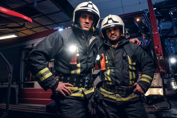 Dos bomberos se abrazan y miran a la cámara de pie cerca del camión de bomberos por la noche en un depósito de incendios — Foto de Stock