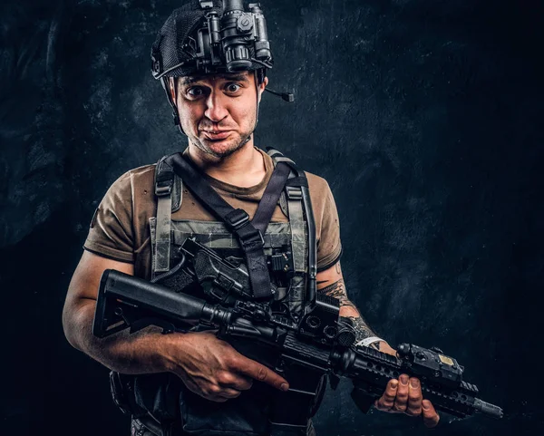 Hombre en camiseta con tatuaje en la mano con armadura corporal y casco con una visión nocturna sosteniendo rifle de asalto y mirando a una cámara con una mirada divertida . — Foto de Stock