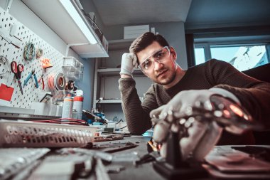 Düşünceli bir bakışla bir kameraya bakarak elektronik teknisyeni gözlük, bir tamir dükkanında bir masa üzerine eğilerek içinde