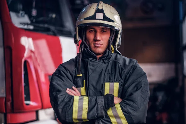 Впевнений пожежник, одягнений в захисну форму, стоїть поруч з пожежним двигуном в гаражі пожежного відділення, схрещує руки і дивиться на камеру — стокове фото