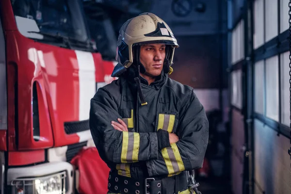 Fireman wearing protective uniform standing next to a fire engine in a garage of a fire department, crossed arms and looking sideways