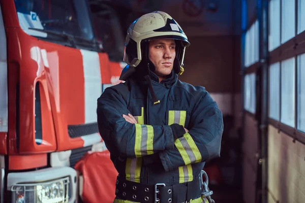 Tűzoltó visel védő egységes mellette egy tűzoltóautó, egy garázs, egy tűzoltóság, karokkal és néz körül — Stock Fotó