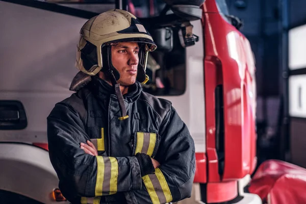 Fireman wearing protective uniform standing next to a fire engine in a garage of a fire department, crossed arms and looking sideways — Stock Photo, Image