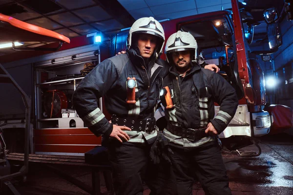 Dos bomberos se abrazan y miran a la cámara de pie cerca del camión de bomberos por la noche en un depósito de incendios — Foto de Stock