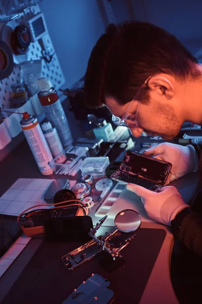 Le réparateur examine attentivement l'intégrité des éléments internes du smartphone dans un atelier de réparation moderne. Illumination avec des lumières rouges et bleues — Photo