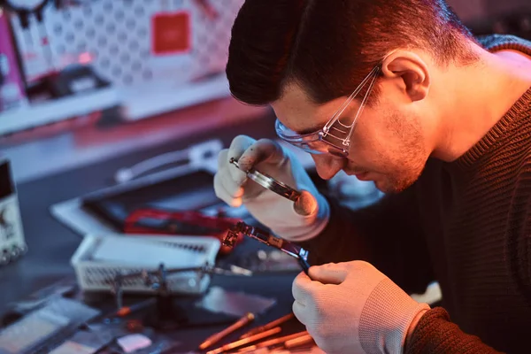 Electronic technician mending a broken tablet carefully examines the chip of the tablet using a magnifying glass. Illumination with red and blue lights