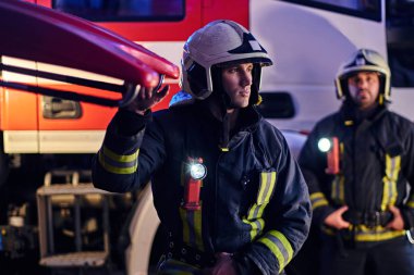 İki cesur itfaiyeci itfaiye kamyonu yanında koruyucu bir üniforma durduğunu giyiyor. Varış gece zaman nöbetçi