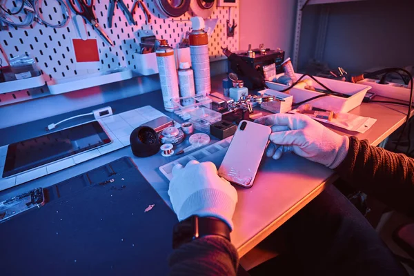 Técnico electrónico sostiene un teléfono inteligente moderno con un cuerpo roto sentado en una mesa en un taller de reparación . —  Fotos de Stock