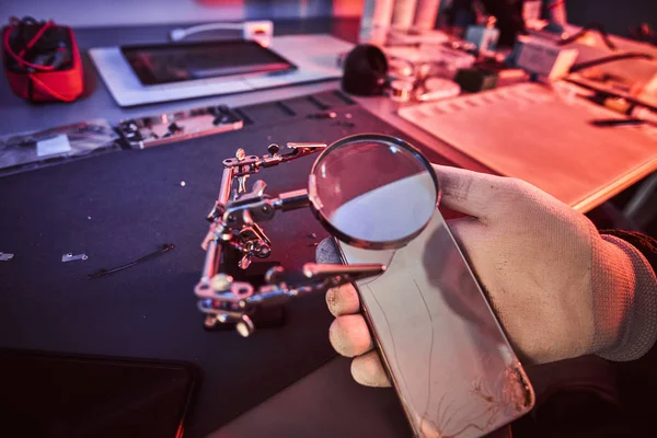 Электронный техник держит современный смартфон со сломанным телом, тщательно исследует повреждения с помощью увеличительного стекла. Освещение красным и синим светом — стоковое фото