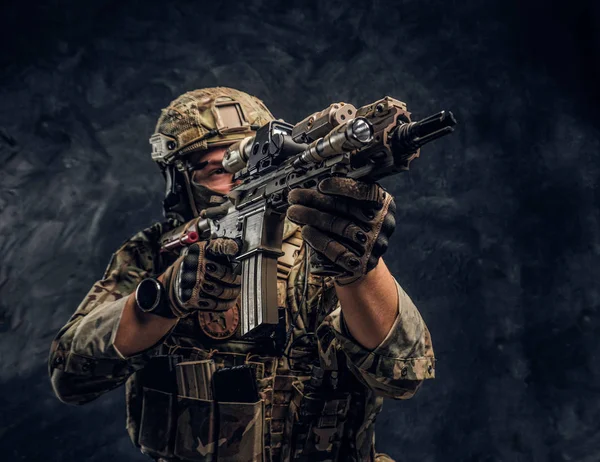 La unidad de élite, soldado de las fuerzas especiales en uniforme de camuflaje que sostiene un rifle de asalto con una mira láser y apunta al objetivo. Foto del estudio contra una pared oscura — Foto de Stock