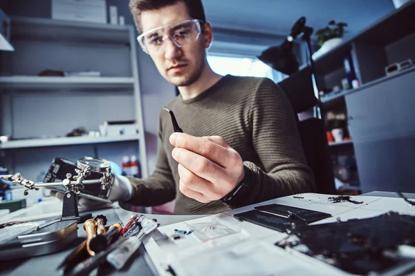 Electronic technician mending a broken phone, looking closely at the little bolt holding it with tweezers
