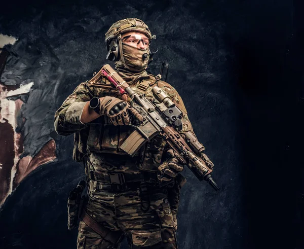 Soldado totalmente equipado con uniforme de camuflaje que sostiene un rifle de asalto. Foto del estudio contra una pared oscura — Foto de Stock