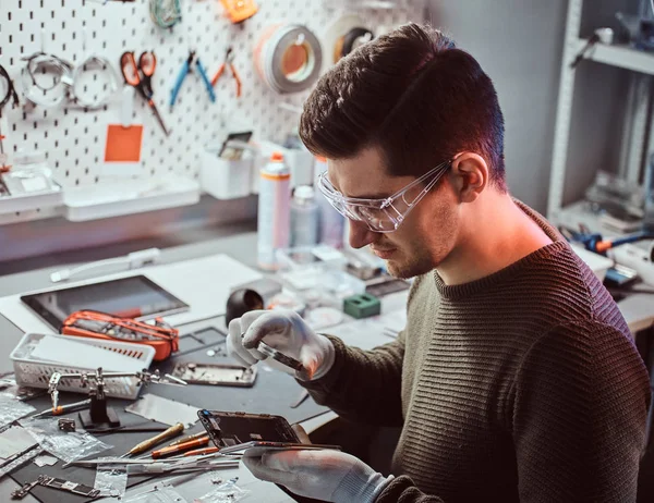 O técnico usa uma lupa para inspecionar cuidadosamente as partes internas do smartphone em uma oficina de reparo moderna — Fotografia de Stock