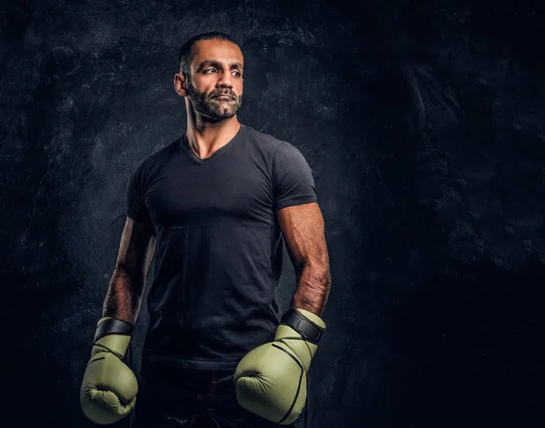 Portrait of a brutal professional fighter in a black shirt and gloves. Studio photo against a dark textured wall — Stock Photo, Image