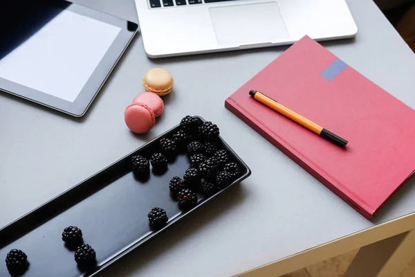 Bacche fresche e macaron francesi colorati, pranzo in ufficio. Laptop, tablet, smartphone e notebook su un tavolo — Foto Stock