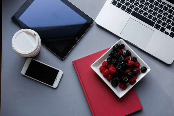 Frambuesas, moras y café, almuerzo saludable en la oficina. Ordenador portátil, tableta, teléfono inteligente y portátil en una mesa — Foto de Stock