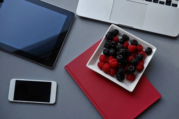 Ordenador portátil, tableta, smartphone y portátil en una mesa. Frambuesas y moras, almuerzo saludable en la oficina — Foto de Stock