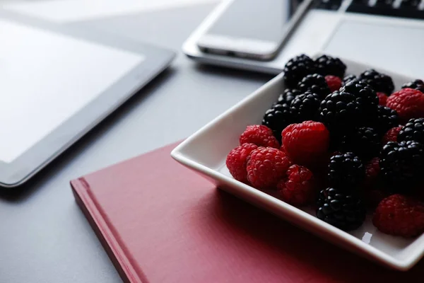 Ordenador portátil, tableta, smartphone y portátil en una mesa. Frambuesas y moras, almuerzo saludable en la oficina — Foto de Stock