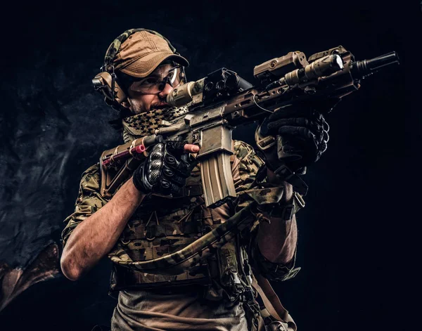 The elite unit, special forces soldier in camouflage uniform holding an assault rifle with a laser sight and aims at the target. Studio photo against a dark wall