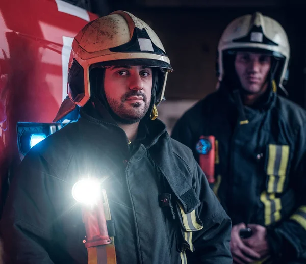 Dos bomberos con uniforme protector de pie junto a un camión de bomberos en un garaje de un departamento de bomberos . — Foto de Stock