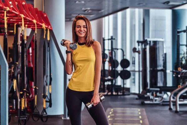 Sorrindo menina fitness fazendo exercícios com halteres, treino no ginásio moderno — Fotografia de Stock