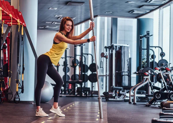 Fitness fille posant pour un appareil photo tout en s'appuyant sur un haltère dans la salle de gym moderne — Photo