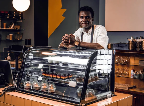 Smiling owner of the coffee shop in uniform looking at camera while leaning on a counter — Stock Photo, Image